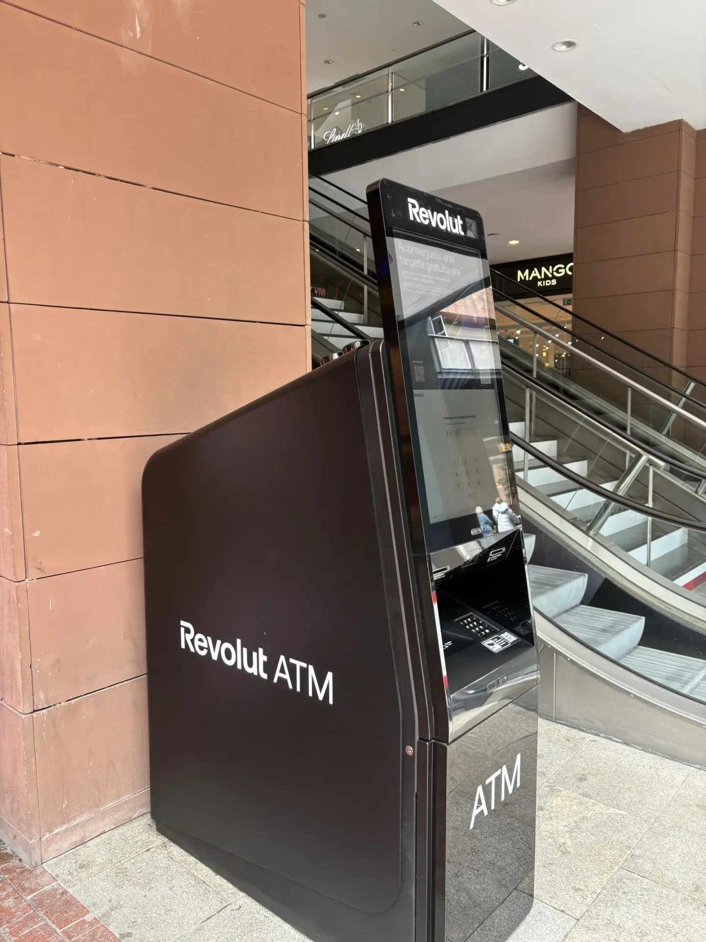 Side angle of the Revolut ATM kiosk, with shopping centre escalator in the background